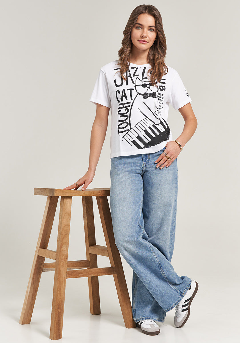 A woman with wavy brown hair stands by a tall wooden stool, wearing the Princess Goes Hollywood T-Shirt Jazz Club Cat, light blue wide-leg jeans, and white sneakers with black stripes. She looks at the camera with a relaxed expression.