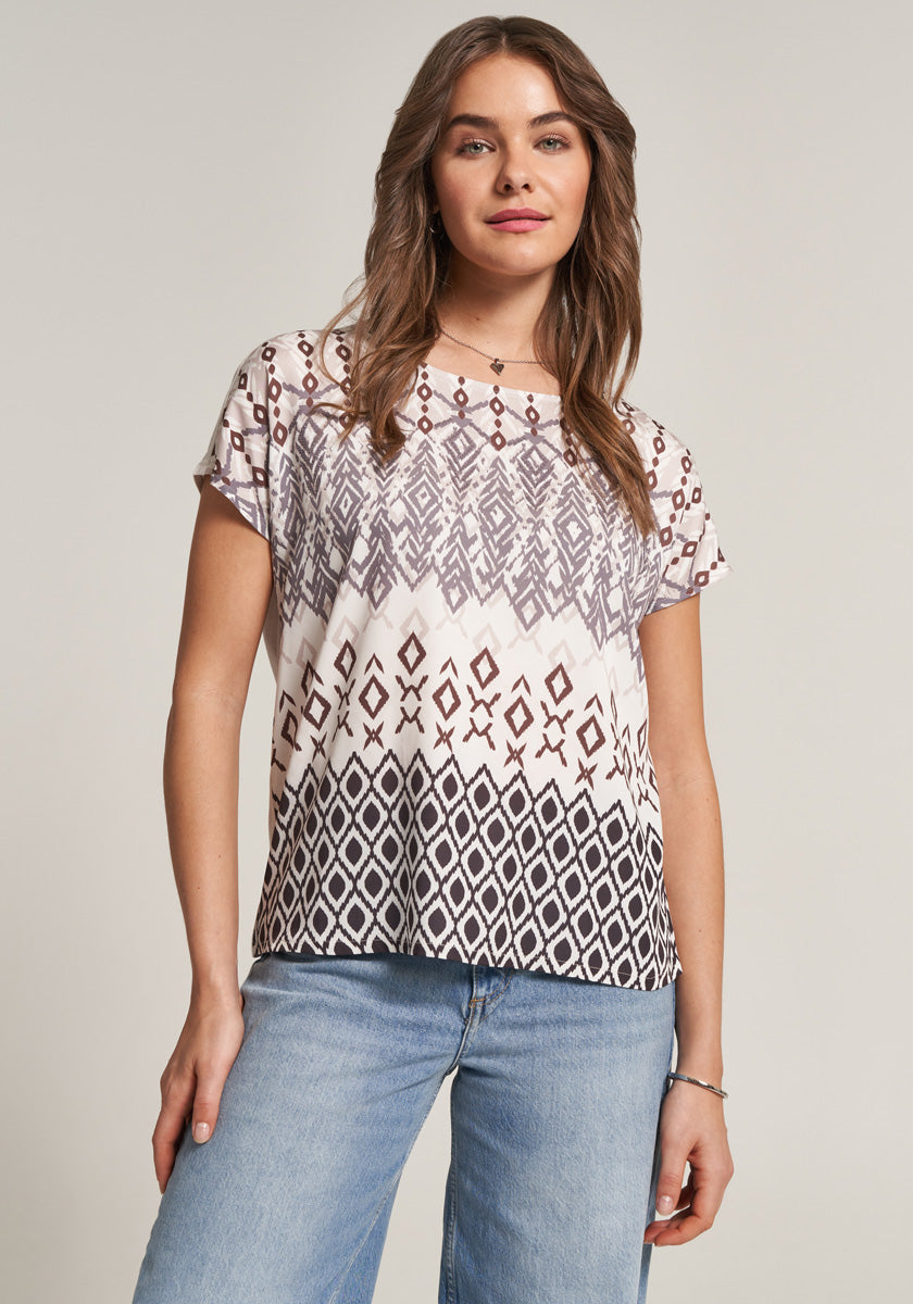 A woman with long brown hair wears a loose, short-sleeved Princess Goes Hollywood T-shirt aus Baumwolle-Modal-Mix mit geometrischem Braun-Schwarz-Print and light blue jeans, facing forward and smiling slightly against a plain background.