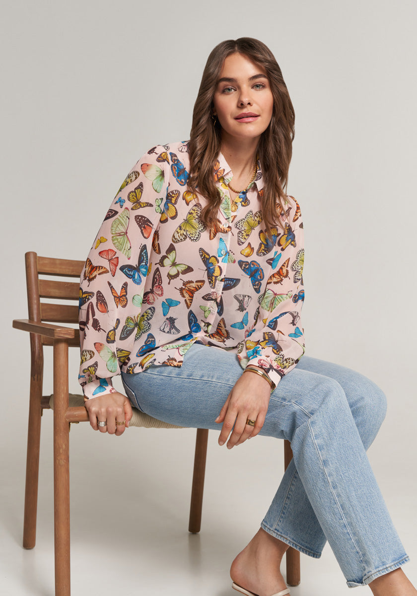 A woman with wavy brown hair sits sideways on a wooden chair, wearing a sheer Princess Goes Hollywood Schmetterling-Print Bluse with Hemdkragen and light blue jeans. She smiles softly at the camera, hands on her lap, against a plain light gray background.