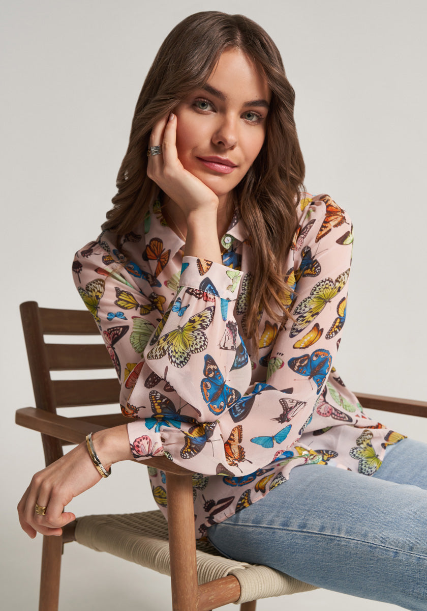 A woman with wavy brown hair sits on a wooden chair, smiling softly. She wears a light pink Princess Goes Hollywood Bluse with Hemdkragen and colorful Schmetterling-Prints, paired with light blue jeans. The background is plain and well-lit.
