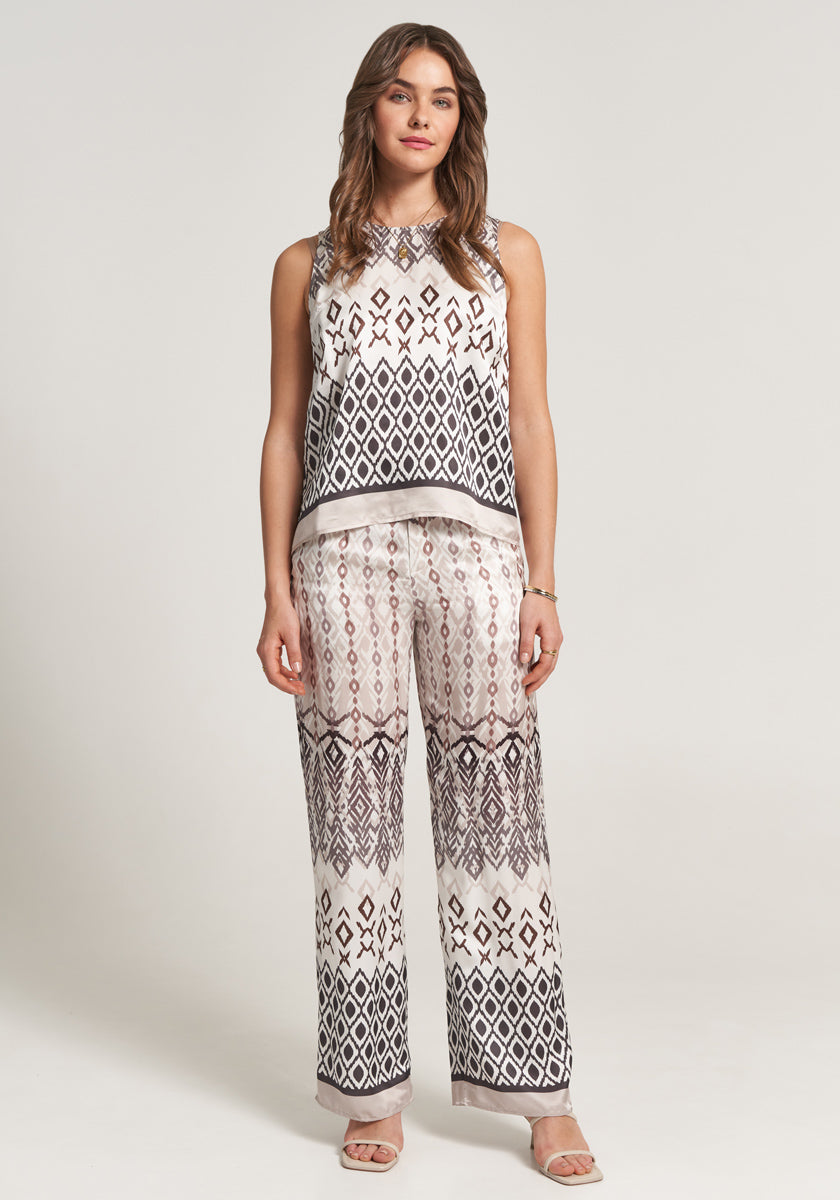 A woman with light wavy hair stands facing forward in a Princess Goes Hollywood blouse with silk blend and wide-leg pants featuring a bold black, white, and taupe geometric print. She wears neutral sandals and is set against a plain, light background.