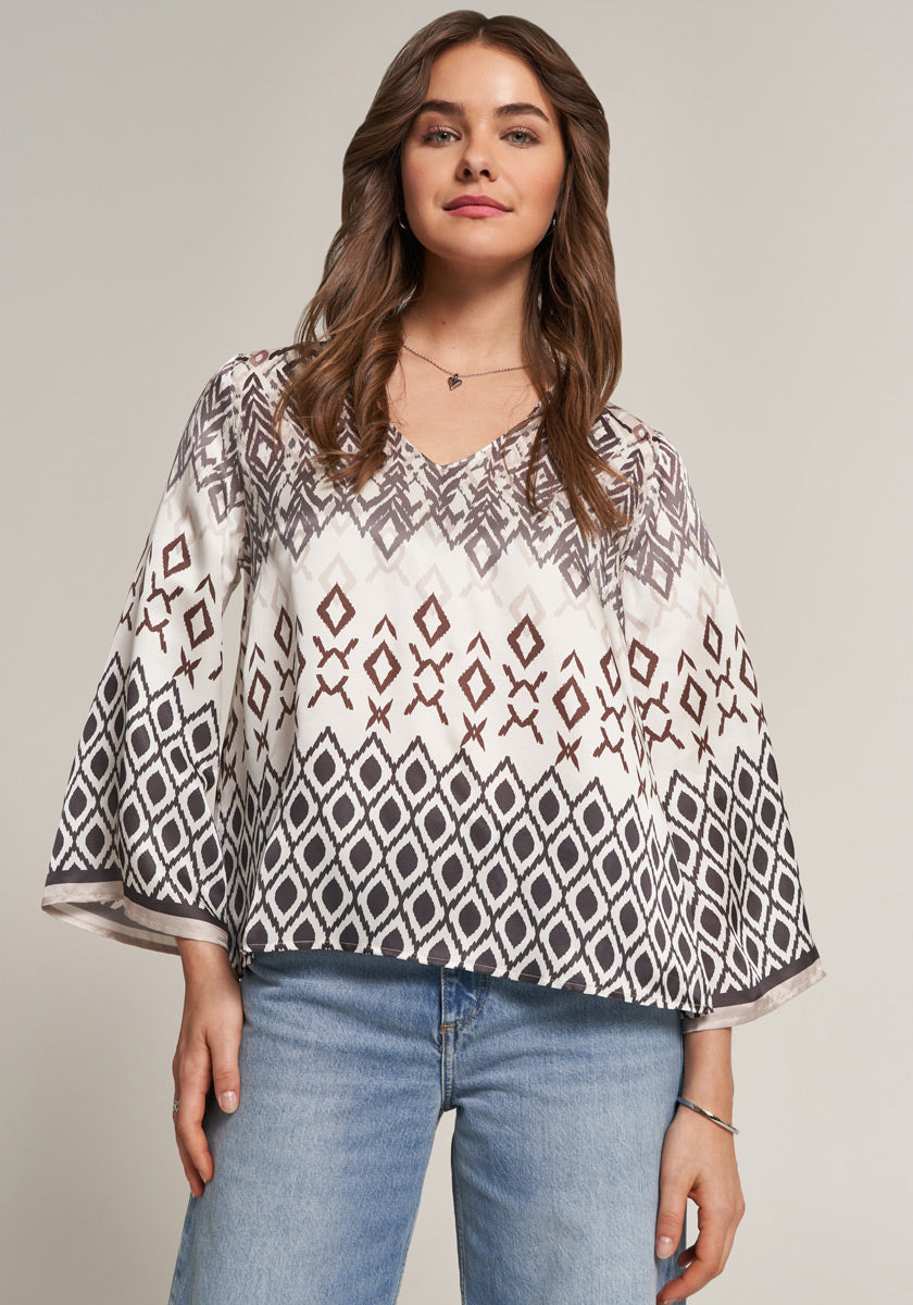 A woman with wavy brown hair wears a loose Princess Goes Hollywood blouse featuring a geometric brown and black print on white. She pairs it with light blue jeans and minimal jewelry, smiling softly against a light background.