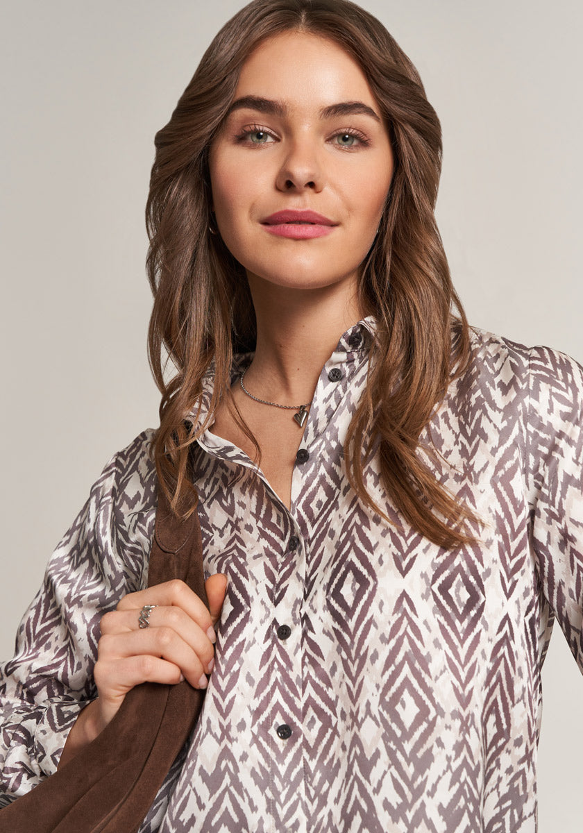 A woman with long brown hair wears a gray and white Princess Goes Hollywood Hemdbluse with geometric print. She has a brown shoulder strap, natural makeup, and looks confidently at the camera with a slight smile.