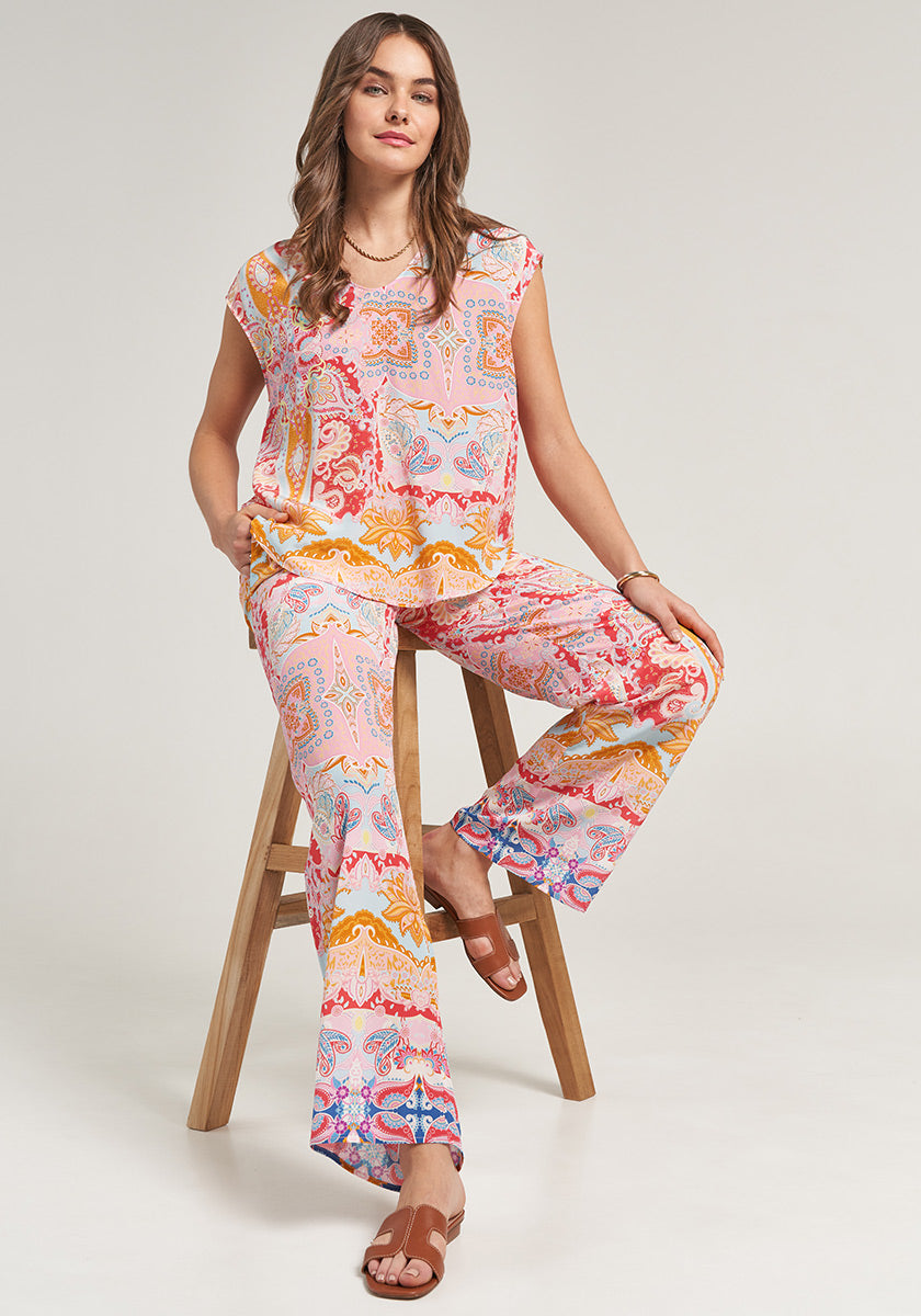 A woman with long brown hair and sandals sits confidently on a wooden stool, wearing a matching allover-paisley short-sleeve top and wide-leg elastic-waist pants by Princess Goes Hollywood, set against a plain light background.