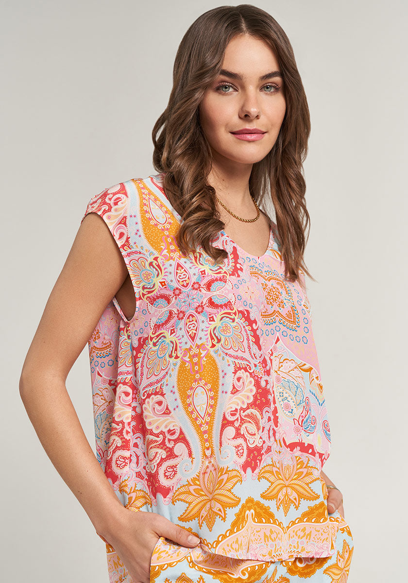 A woman with loose brown hair wears a colorful paisley V-neck Seidenbluse by Princess Goes Hollywood in pink, orange, red, and yellow. She stands sideways and smiles softly with one hand in her pocket against a plain light background.