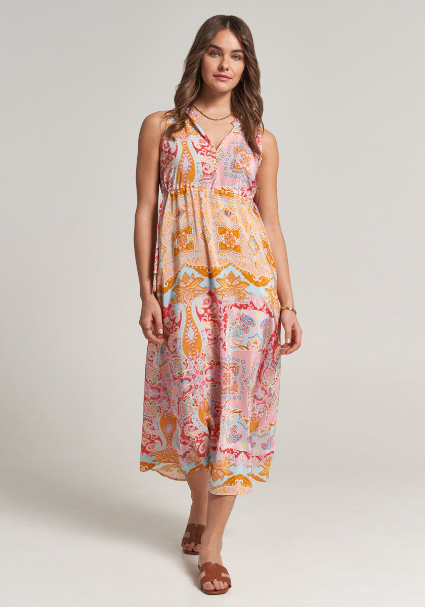 A woman poses against a plain light background wearing a sleeveless Princess Goes Hollywood Seidenkleid with V-neck and an allover print in pink, orange, and yellow paisley and geometric patterns. She accessorizes with wavy hair, brown flat sandals, and a necklace.