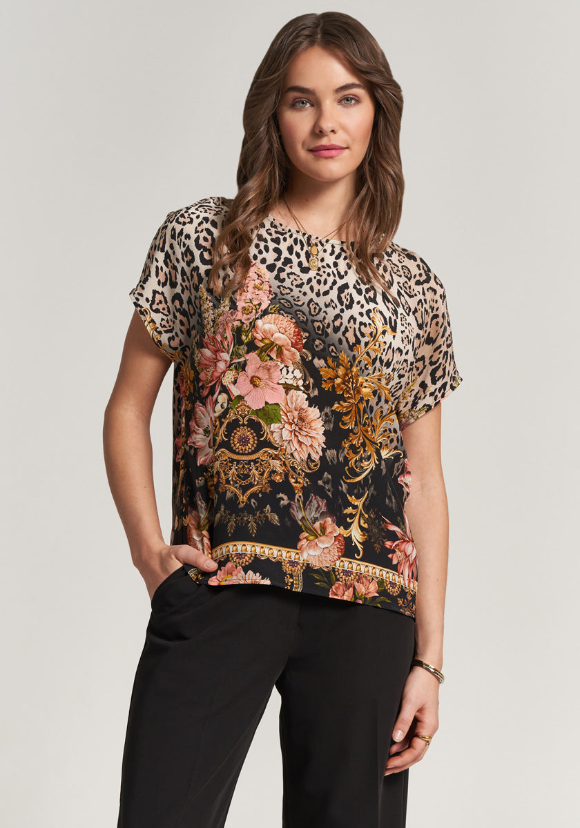 A woman with wavy brown hair smiles softly, one hand in her black pants pocket, wearing a Princess Goes Hollywood silk blouse with a floral leopard print in beige, black, pink, and gold against a plain light background.