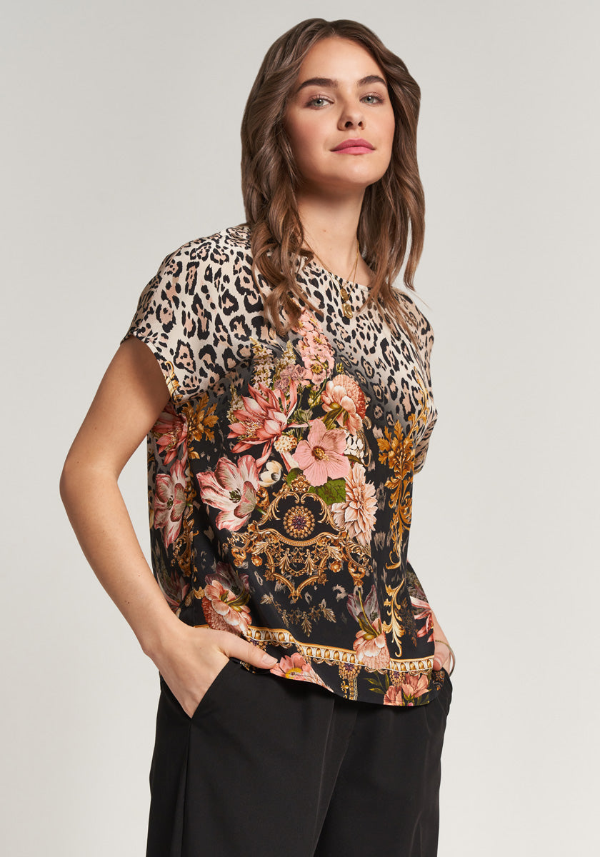 A woman poses confidently with one hand in her pocket, wearing a Princess Goes Hollywood Seide Bluse adorned with floral leopard print on the shoulders and ornate pink, gold, and black designs. Her wavy brown hair frames her face as she looks at the camera.