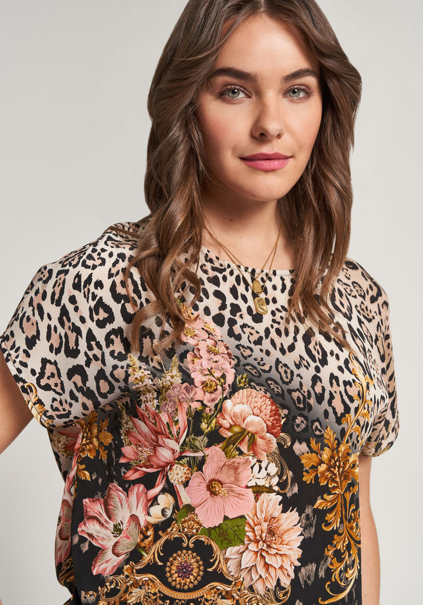 A woman with wavy brown hair, fair skin, and green eyes wears a Princess Goes Hollywood Seidenbluse with floraler Leoprint in pink, peach, and yellow. She gazes confidently at the camera, her gold necklace adding elegance to her look.