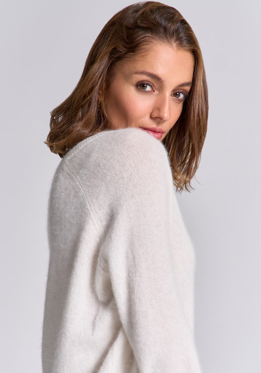 A woman with wavy, shoulder-length brown hair wears a soft Princess Goes Hollywood Strickpullover mit Wolleanteil. She turns slightly, looking over her shoulder with a gentle expression against a plain light gray background.