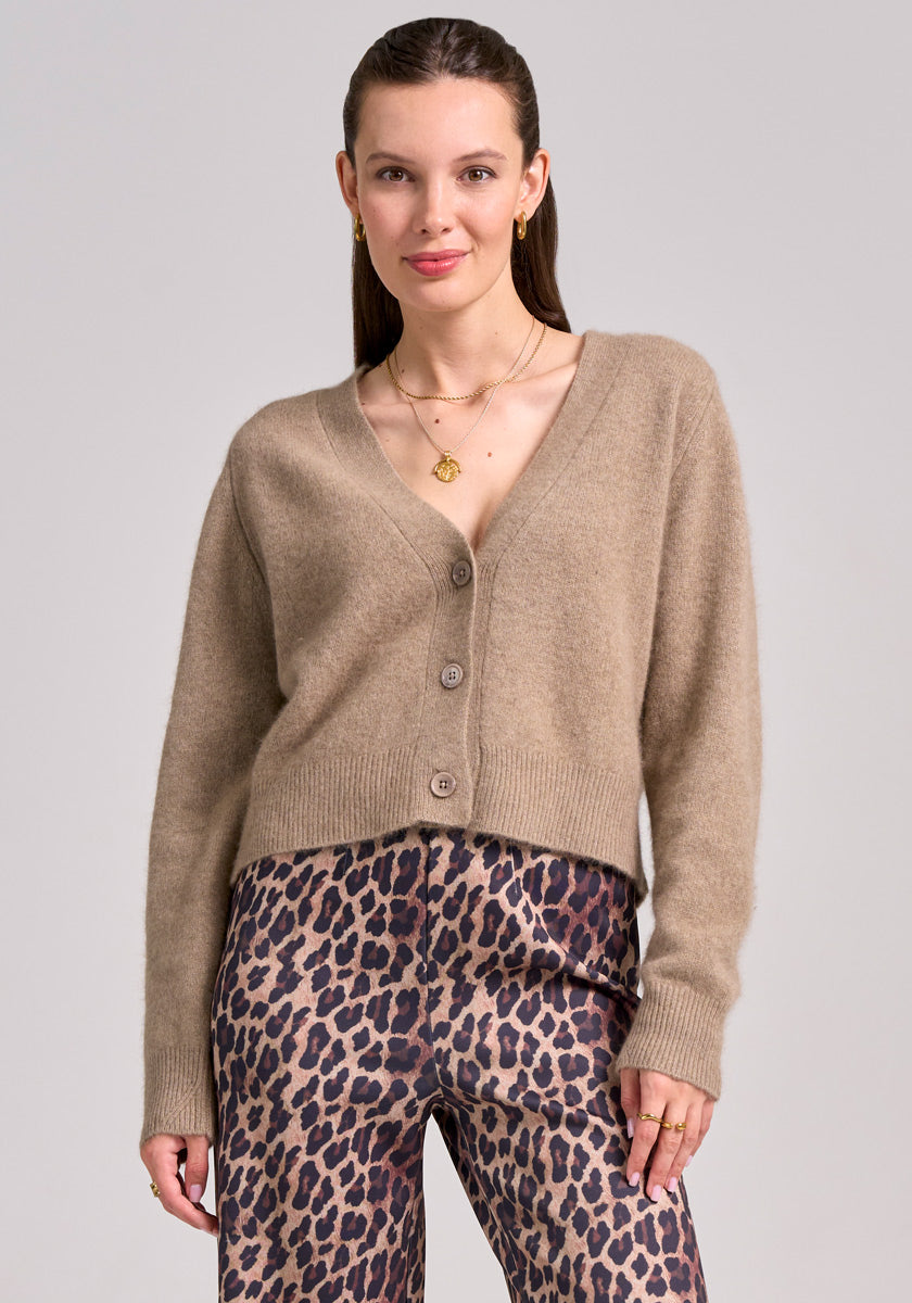 A woman with straight brown hair wears a beige Princess Goes Hollywood cardigan with V-neck and wool, paired with high-waisted, loose-fitting leopard print pants. Layered gold necklaces and rings complete her look as she smiles against a light gray background.