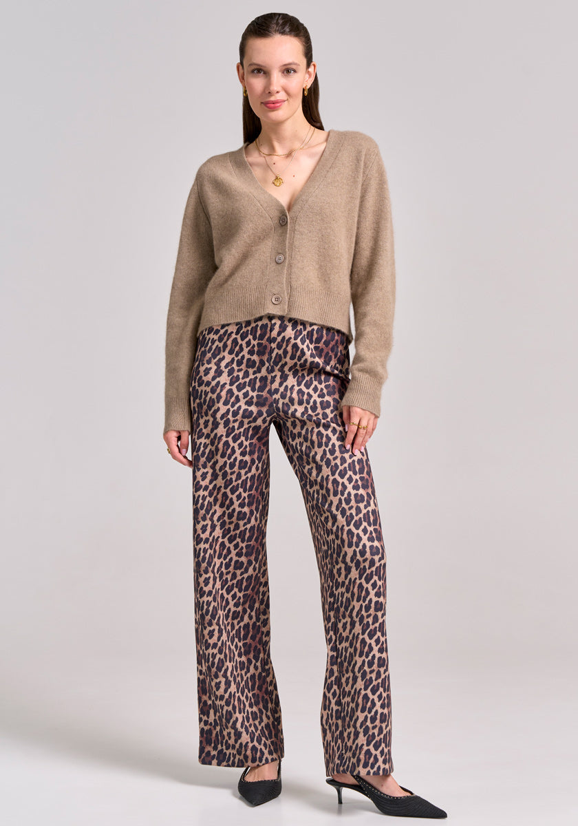 A woman stands against a light gray background wearing a tan V-Ausschnitt Strickjacke mit Wolleanteil from Princess Goes Hollywood, leopard print wide-leg pants, black pointed-toe heels, and a gold necklace. Her straight brown hair is tucked behind her ears.