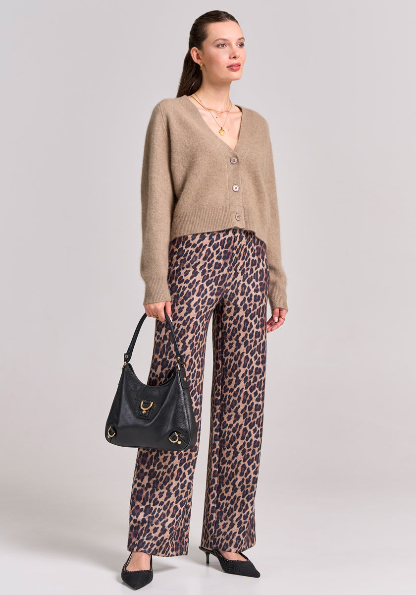 A woman stands against a light gray background wearing a tan V-neck Strickjacke by Princess Goes Hollywood, brown leopard-print wide-leg pants, black pointed heels, and a black shoulder bag, accessorized with gold necklaces and small hoop earrings.