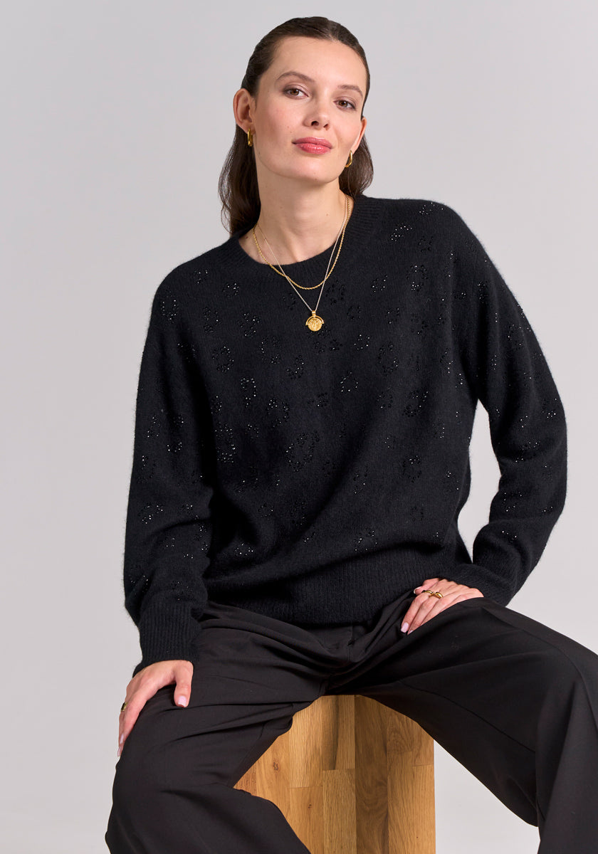 A woman sits on a wooden stool against a light gray background. She wears a black Princess Goes Hollywood Strickpullover mit Wolleanteil, wide-leg black pants, gold necklaces, and small earrings. Her dark hair is slicked back as she looks calmly at the camera.