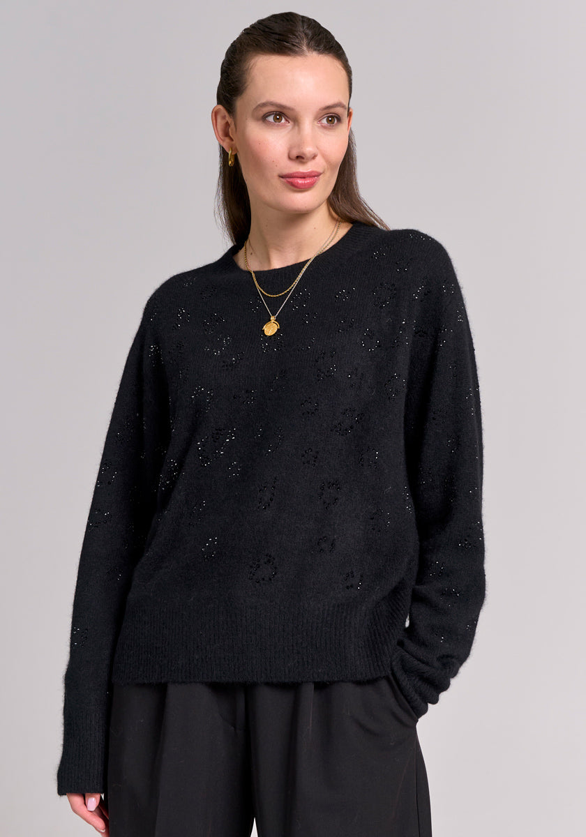 A woman with straight brown hair, tucked behind her ears, wears a schwarzer Strickpullover mit Wolleanteil by Princess Goes Hollywood and loose black pants. She accessorizes with small gold hoops and layered necklaces against a light gray background.