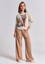 A woman with shoulder-length brown hair smiles confidently in a beige Princess Goes Hollywood Strickpullover aus Merino-Alpaka-Mix featuring a large Waschbär-Motiv, paired with loose tan trousers and white sneakers against a plain background.