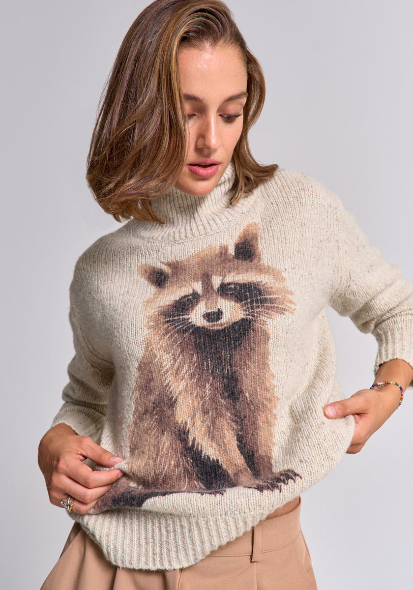 A woman with light brown hair gently touches her cream Princess Goes Hollywood Merino-Alpaka-Mix sweater, which features a large, realistic Waschbär motif on the front, while looking down and resting her other hand on her hip.