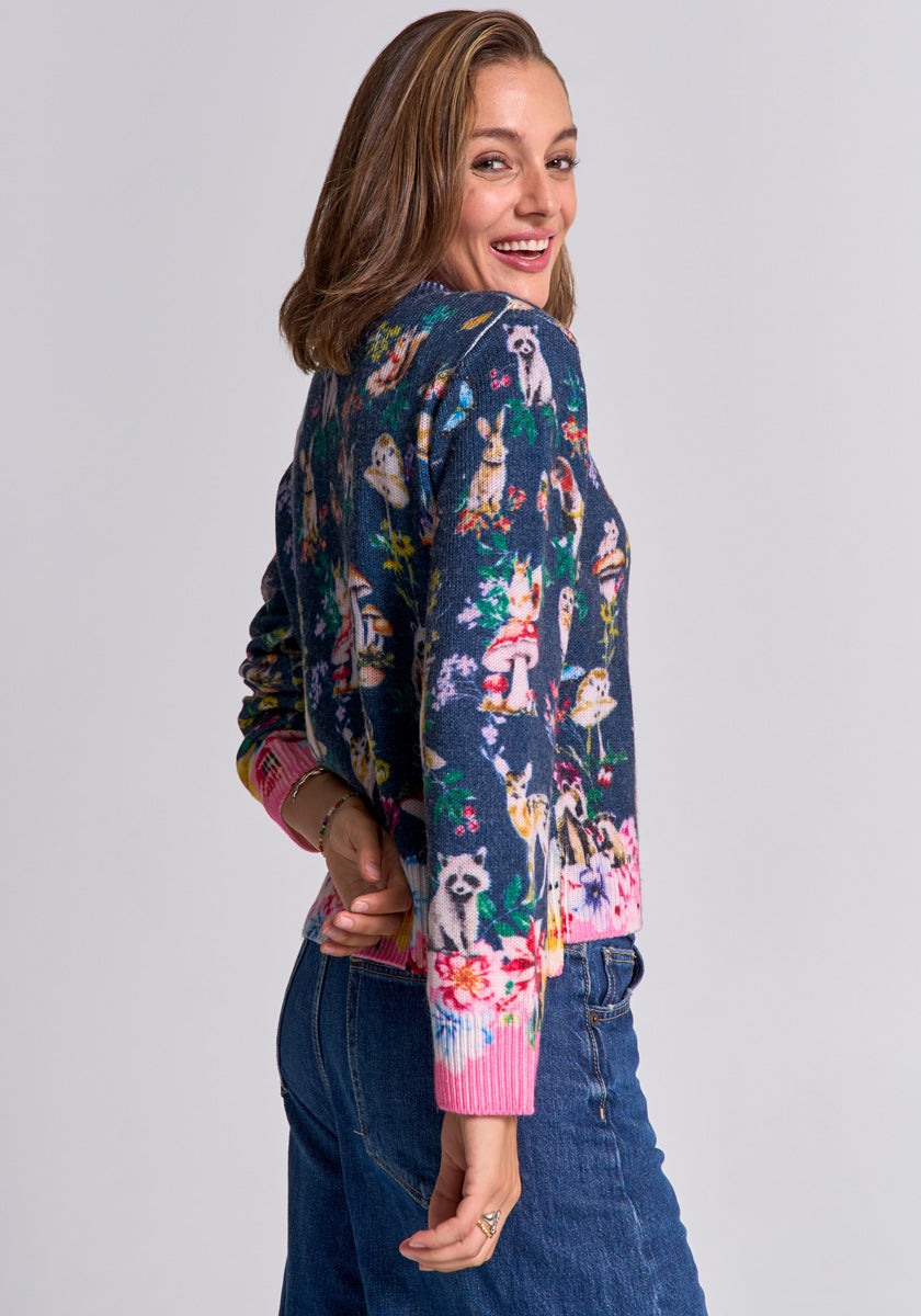 A smiling woman with shoulder-length brown hair wears a colorful Princess Goes Hollywood Strickpullover featuring dog illustrations and pink cuffs, paired with blue jeans, turning slightly as she looks over her shoulder against a plain light background.