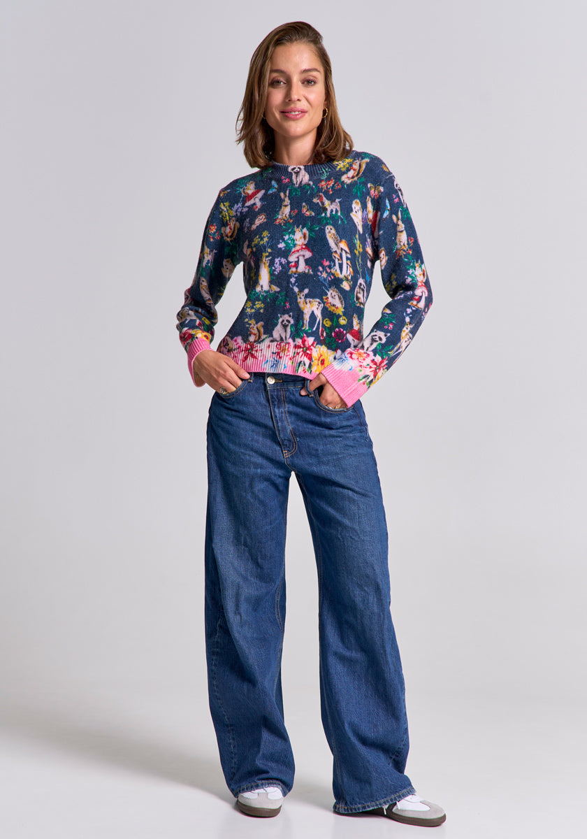 A woman smiles with hands in her pockets, wearing a colorful Princess Goes Hollywood Merinowolle Pullover with pink cuffs and hem, wide-leg blue jeans, and white sneakers. Her shoulder-length brown hair frames her face against a light gray background.