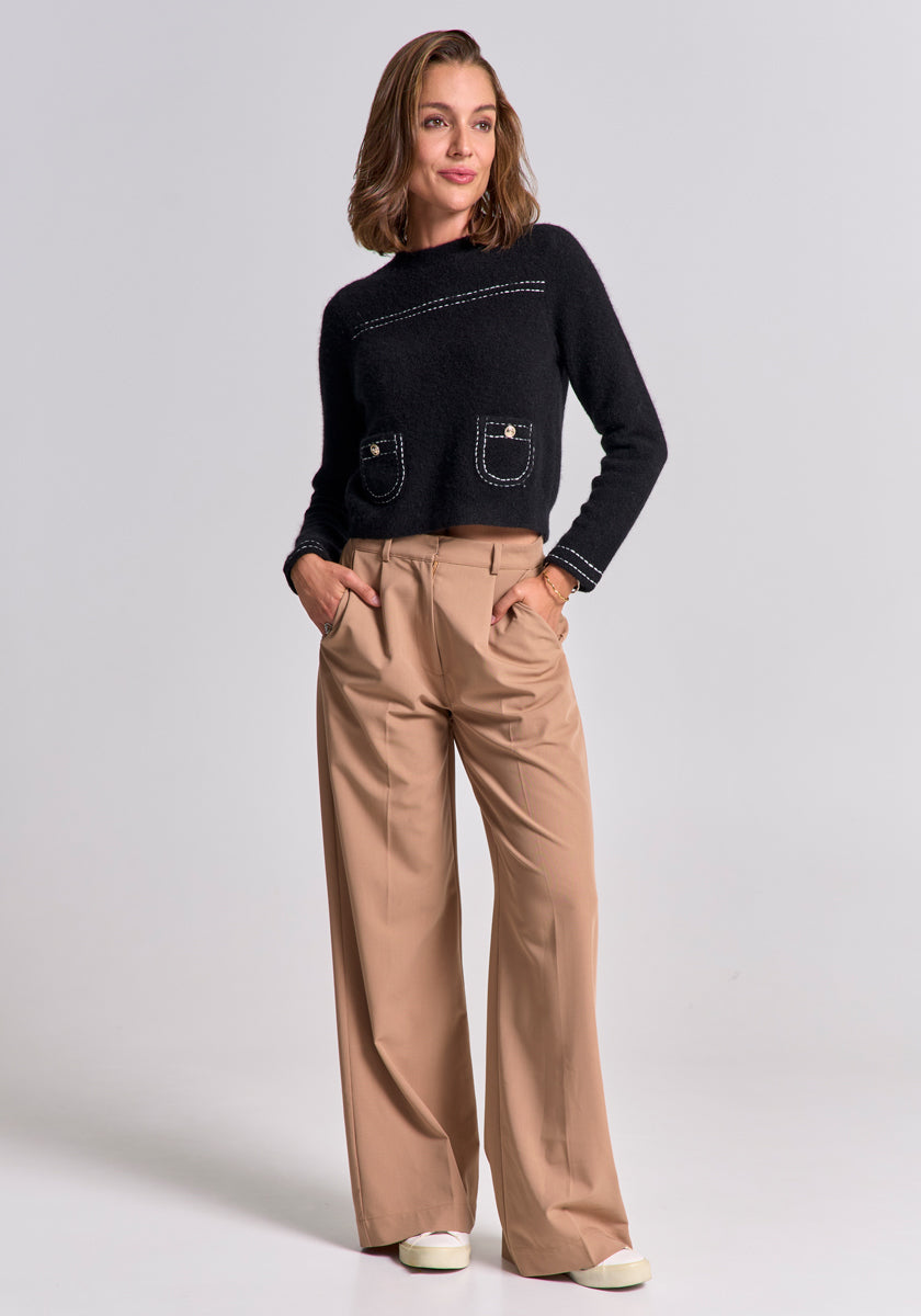 A woman with shoulder-length brown hair smiles in a relaxed pose, wearing beige wide-leg trousers and a black Merino-Kaschmir-Pullover mit Rundhalsausschnitt by Princess Goes Hollywood, hands in pockets and white shoes, against a plain background.