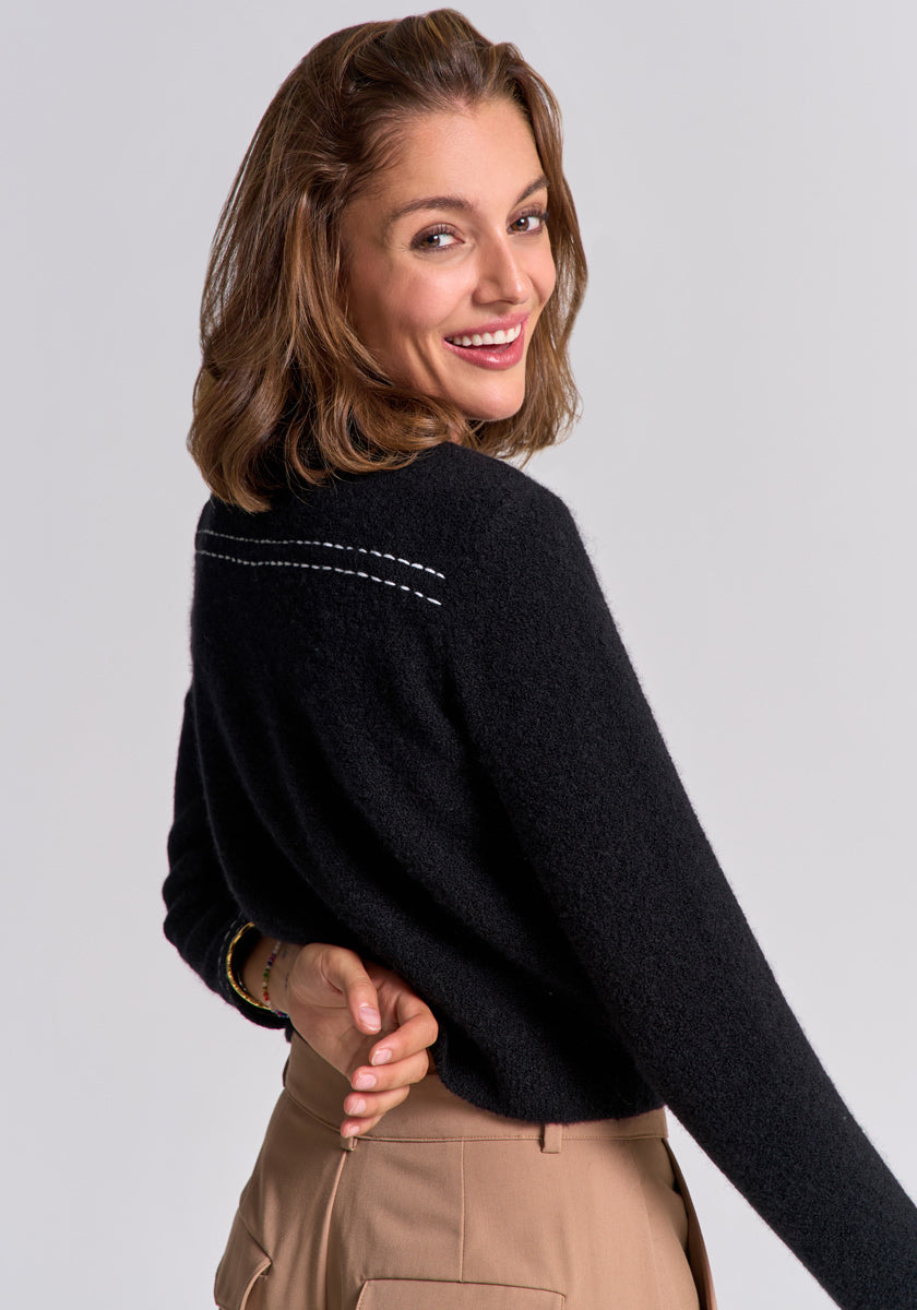 A woman with shoulder-length brown hair smiles over her shoulder, wearing a Princess Goes Hollywood black Merino-cashmere sweater with white embroidery on the back and high-waisted beige pants against a plain light gray background.
