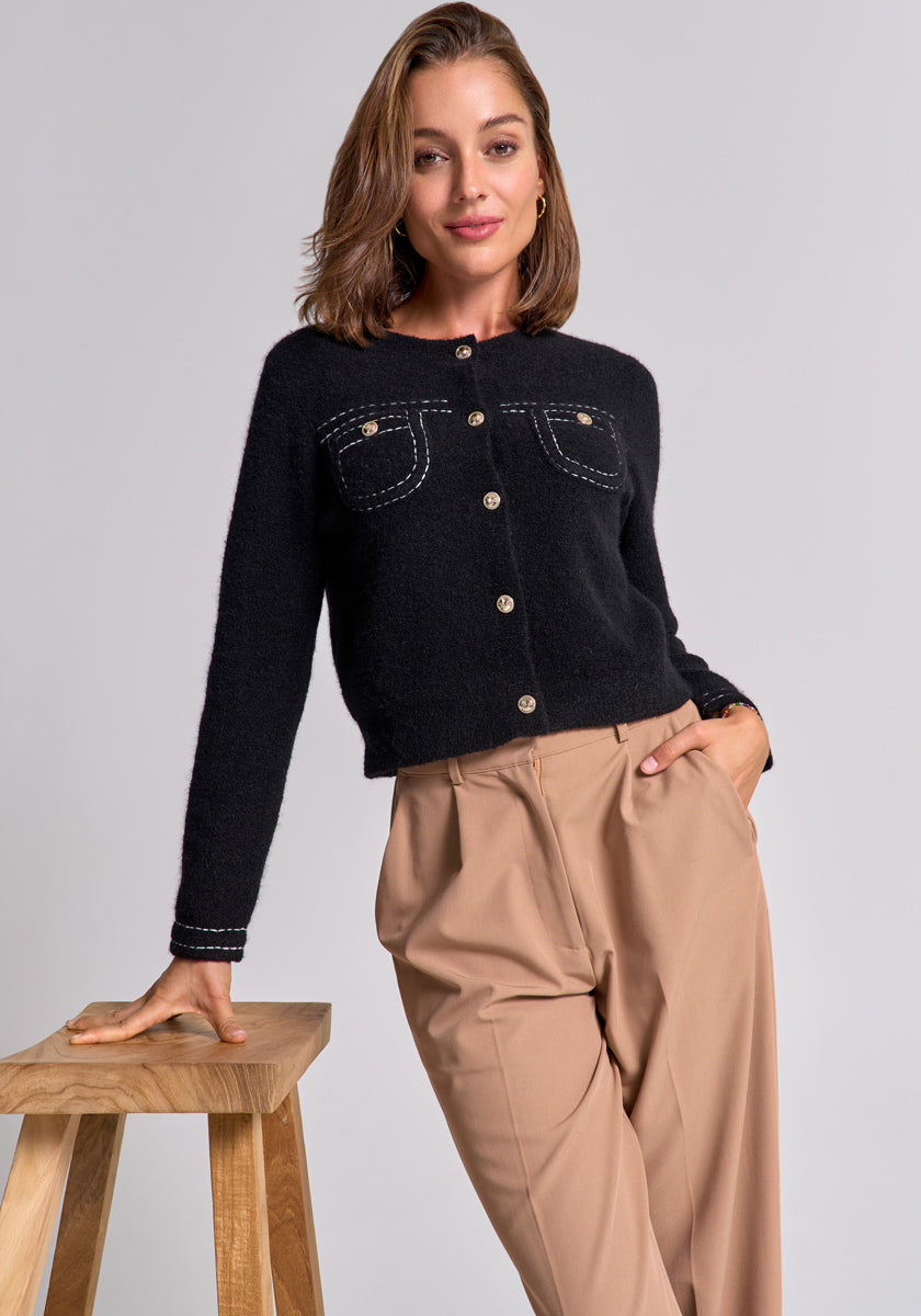 A woman with shoulder-length brown hair wears a Princess Goes Hollywood Merino-Kaschmir-Strickjacke with gold buttons and front pockets, paired with high-waisted beige trousers. She leans on a wooden stool, smiling, against a light gray background.