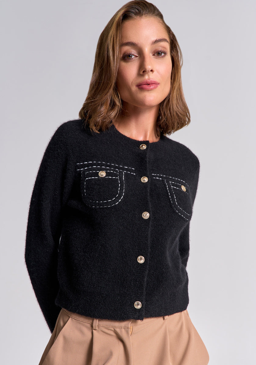 Eine Frau mit schulterlangem, hellbraunem Haar trägt von Princess Goes Hollywood eine schwarze Merino-Kaschmir-Strickjacke mit goldfarbenen Knöpfen und Rundhalsausschnitt sowie beigefarbene, plissierte High-Waist-Hosen.