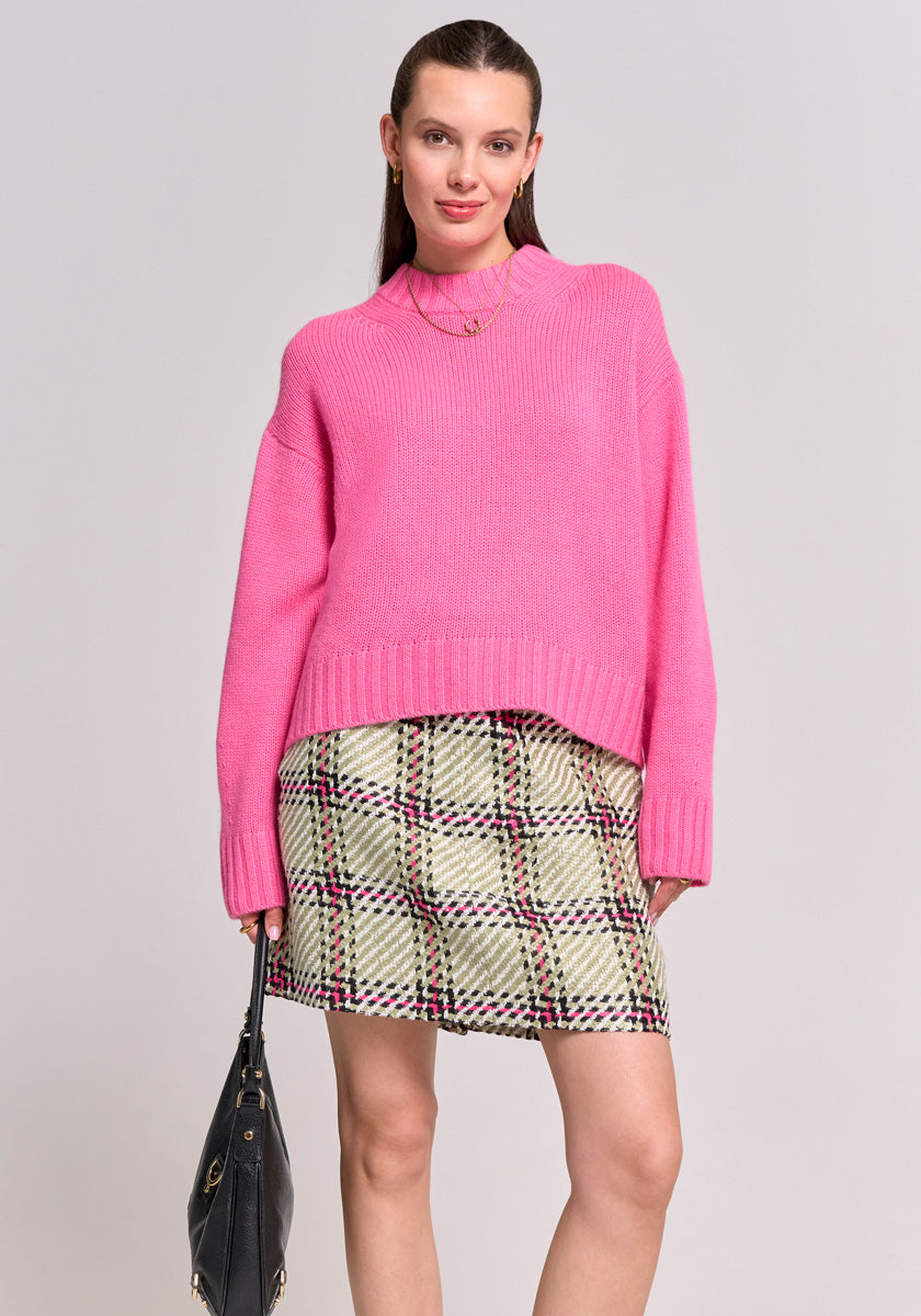 A woman faces forward in a bright pink Princess Goes Hollywood Damen Stehkragen Kaschmir Pullover and green-white plaid mini skirt. She carries a black handbag, her straight brown hair tucked behind her ears. The background is plain and light.