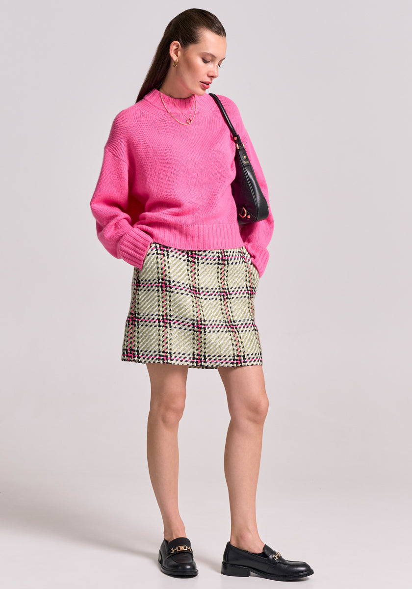 A woman with straight brown hair wears a bright pink Princess Goes Hollywood Kaschmir Pullover mit Stehkragen, a cream plaid mini skirt with black and red lines, black loafers, gold jewelry, and carries a small black shoulder bag against a plain background.