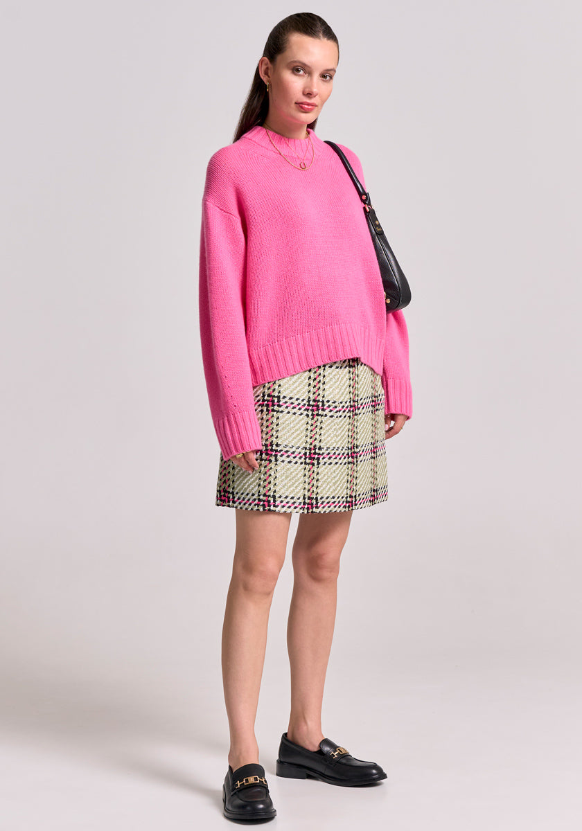 A woman poses against a light gray background wearing a bright pink Princess Goes Hollywood Kaschmir Pullover mit Stehkragen, a green-black-white plaid mini skirt, black loafers with gold accents, and a black shoulder bag—a chic Damen look.