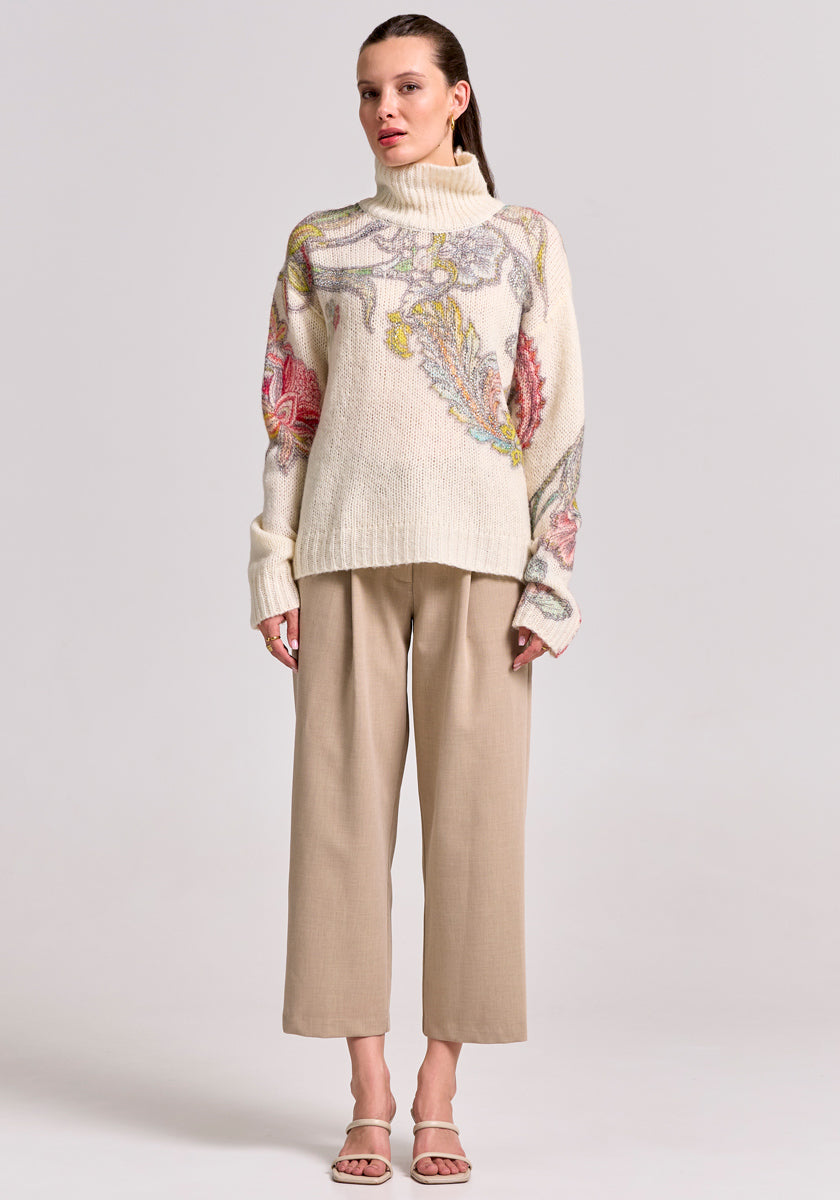 A woman faces forward in a cream Princess Goes Hollywood Rollkragen Wolle-Alpaka-Mix Pullover with colorful abstract floral patterns, styled with beige cropped trousers and white open-toe heels. Her slicked-back hair contrasts against a light gray background.