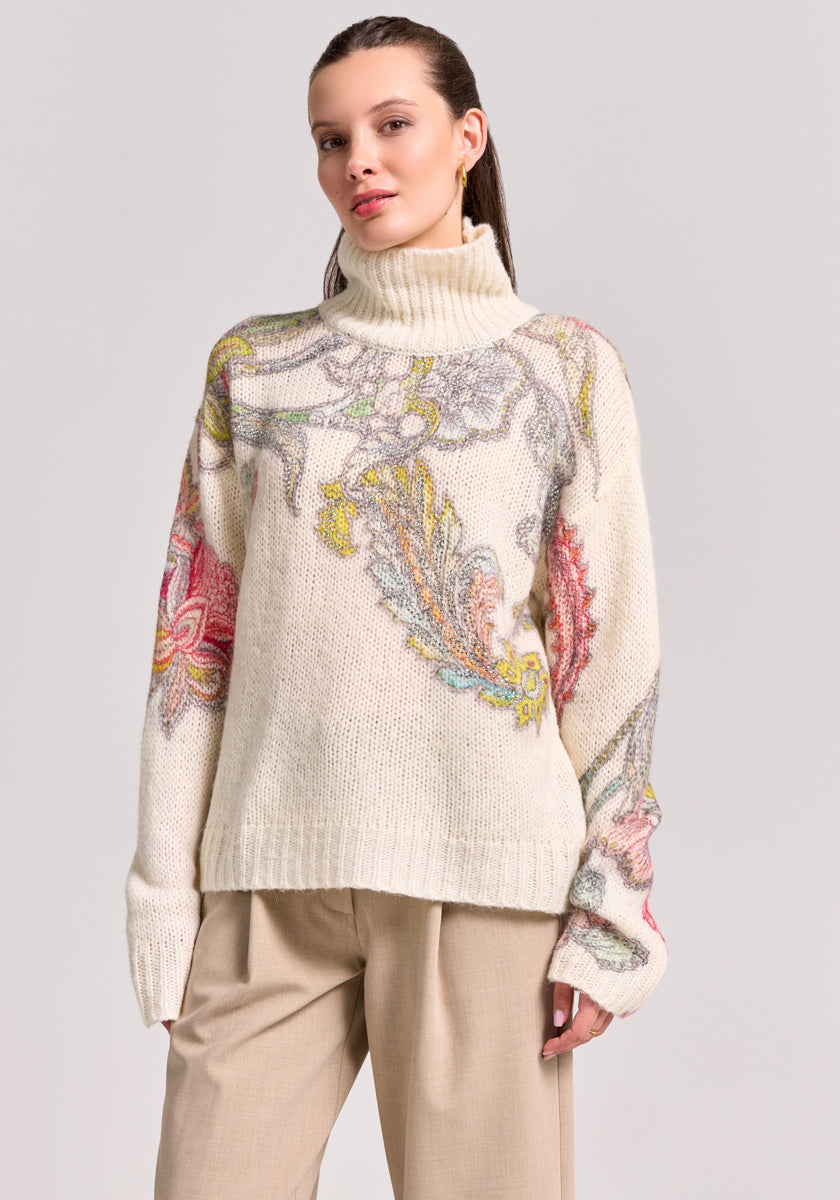 A light-skinned woman with brown hair pulled back models a Princess Goes Hollywood Wolle-Alpaka-Mix Rollkragen-Pullover, showcasing colorful floral and paisley motifs. She pairs it with light beige wide-leg pants against a plain gray backdrop.