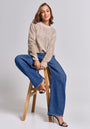 A smiling woman with shoulder-length brown hair sits on a wooden stool against a gray background, wearing a beige Princess Goes Hollywood Strickpullover Damen, wide-leg blue jeans, and black high-heeled sandals, exuding relaxed confidence.