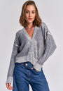 A woman with chin-length brown hair models a grey Merino-Kaschmir-Strickjacke by Princess Goes Hollywood featuring subtle white details, paired with blue high-waisted jeans. She is relaxed, one hand in her pocket, and wears a silver necklace.