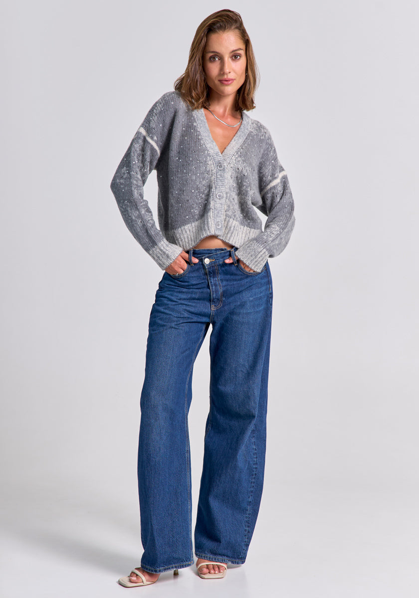 Eine Frau mit schulterlangem braunem Haar trägt eine lockere Strickjacke aus Merino-Kaschmir-Mix von Princess Goes Hollywood über einem weißen Top, Wide-Leg-Jeans und weißen offenen Sandalen vor hellem Hintergrund.