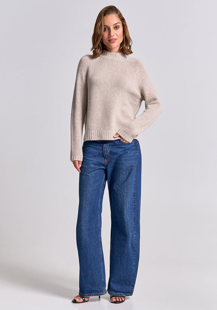 A woman with shoulder-length brown hair and a slight smile wears a beige Princess Goes Hollywood wool-cashmere blend Strickpullover with Stehkragen, loose blue jeans, and black open-toed heels against a plain light gray background.