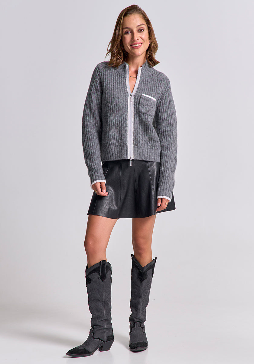 A woman smiles in a gray Princess Goes Hollywood Wolle-Kaschmir-Mix Strickjacke with white pocket detail, paired with a short black leather skirt and knee-high cowboy boots, posing casually against a light background.