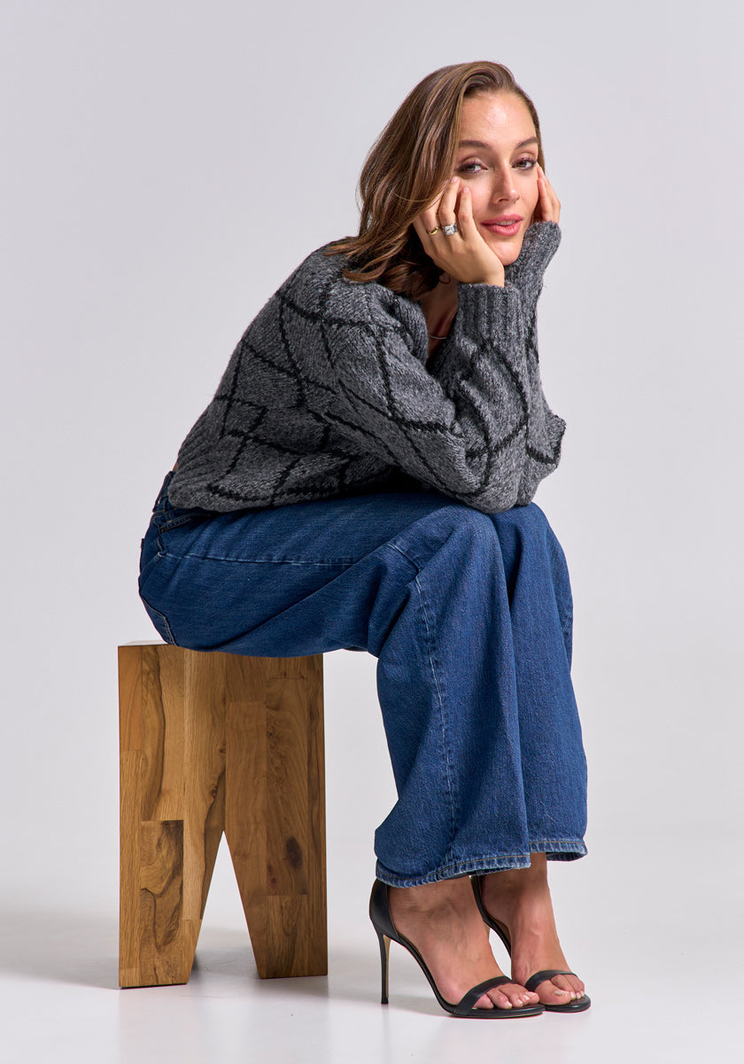 A woman with light brown hair sits on a wooden stool, wearing a loose Princess Goes Hollywood Strickjacke mit Argyle-Muster, wide-leg blue jeans, and black high-heeled sandals. She rests her chin on her hand and looks at the camera, set against a plain light background.