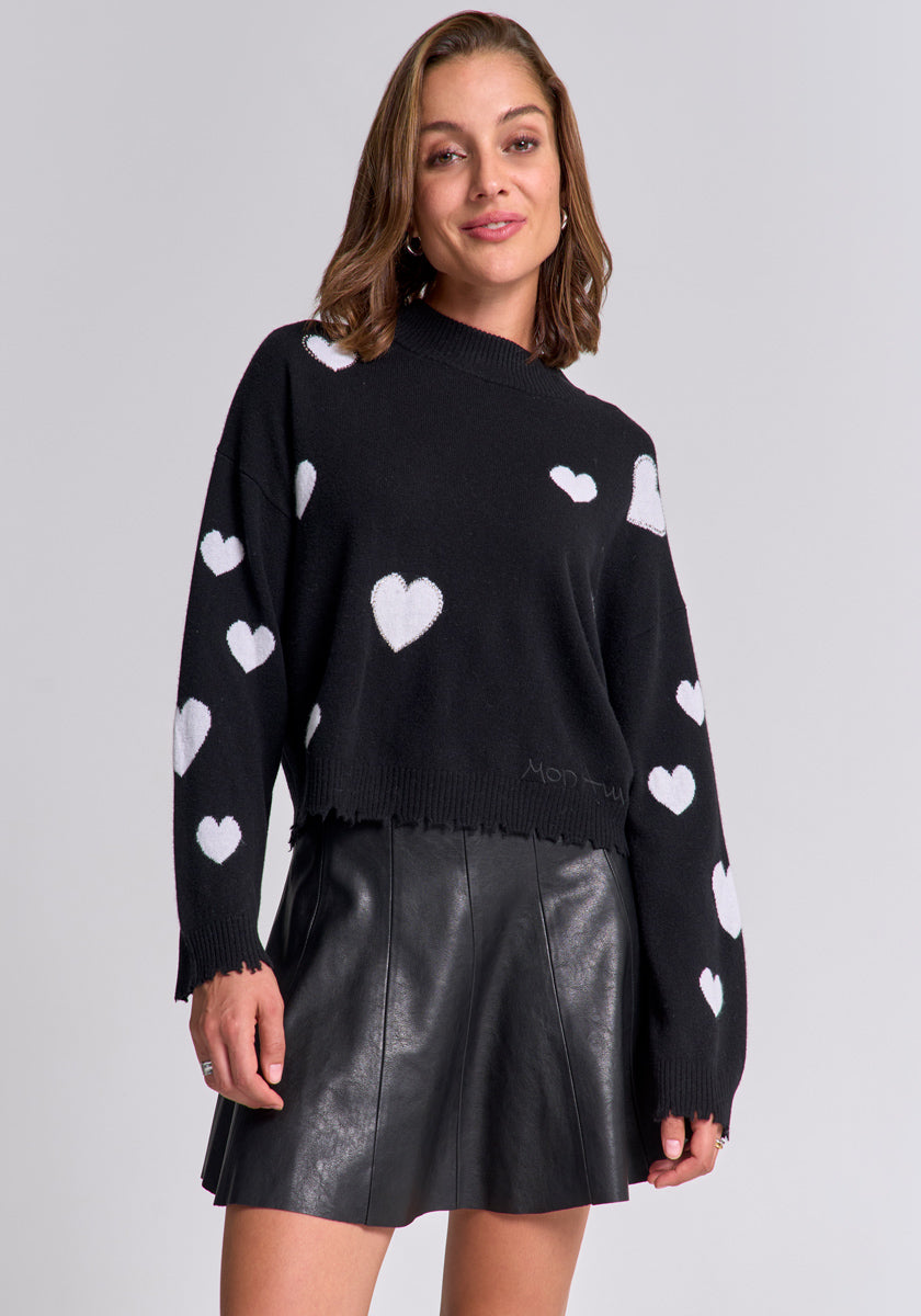 A woman with light brown hair smiles, wearing a Princess Goes Hollywood Feinstrickpullover mit Herz-Muster and a black, high-waisted, pleated leather mini skirt, standing confidently against a plain light gray background.