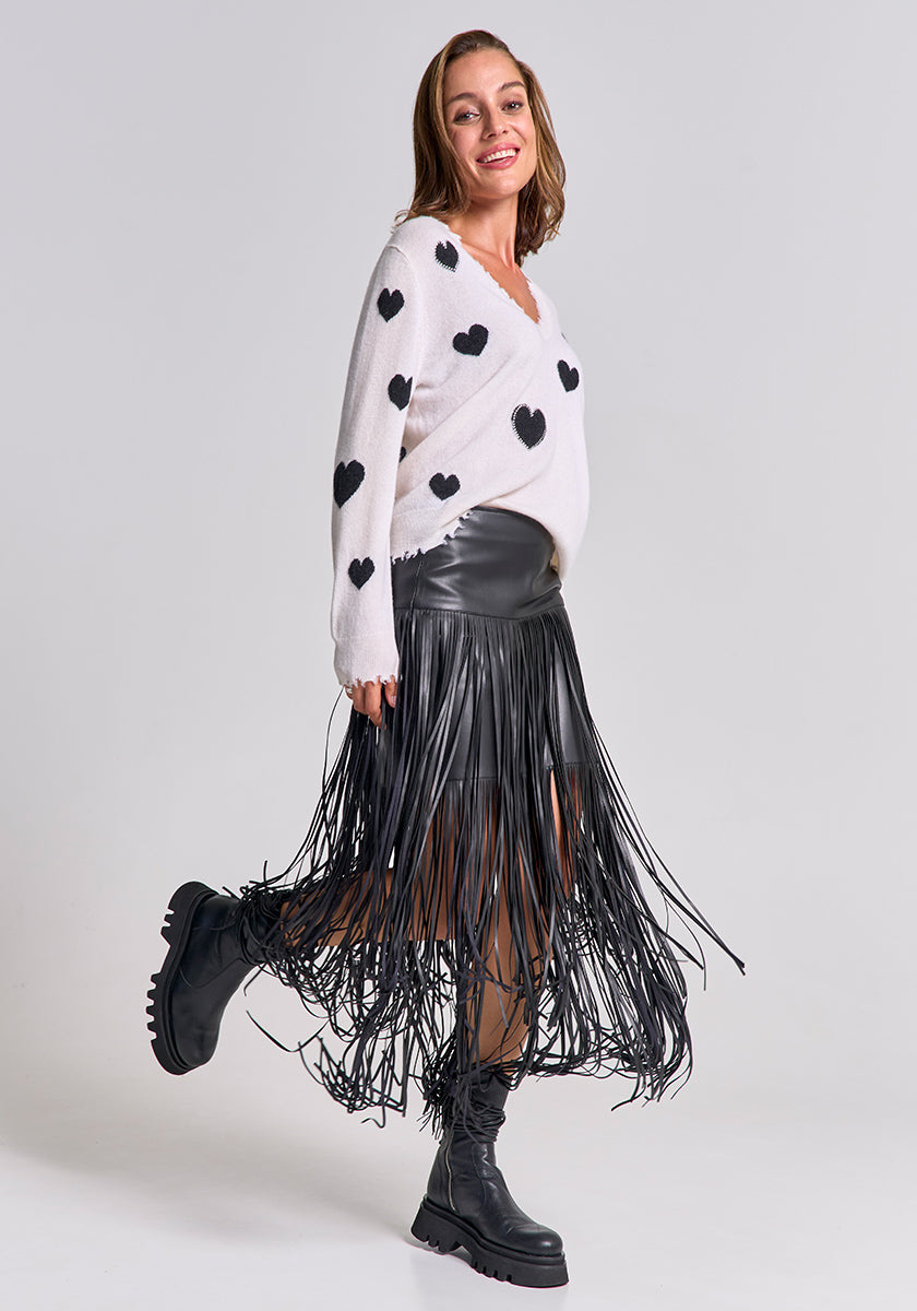 A smiling woman poses in a white sweater with black heart prints, a fringed faux leather mini skirt, and chunky boots from Princess Goes Hollywood against a light grey background, looking confidently at the camera.