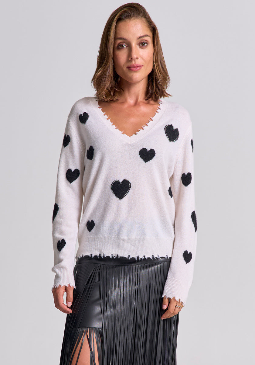 A woman with shoulder-length brown hair wears a white Princess Goes Hollywood wool-cashmere blend pullover with black heart motifs and a black fringed leather skirt, standing against a plain gray background and smiling softly.