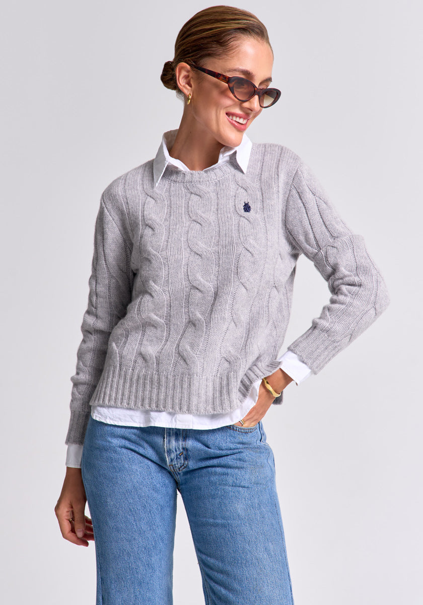 A woman with slicked-back hair wears dark sunglasses, blue jeans, and a light gray Zopfstrick Pullover by Princess Goes Hollywood over a white collared shirt. Smiling, she poses with one hand in her pocket against a light gray background.