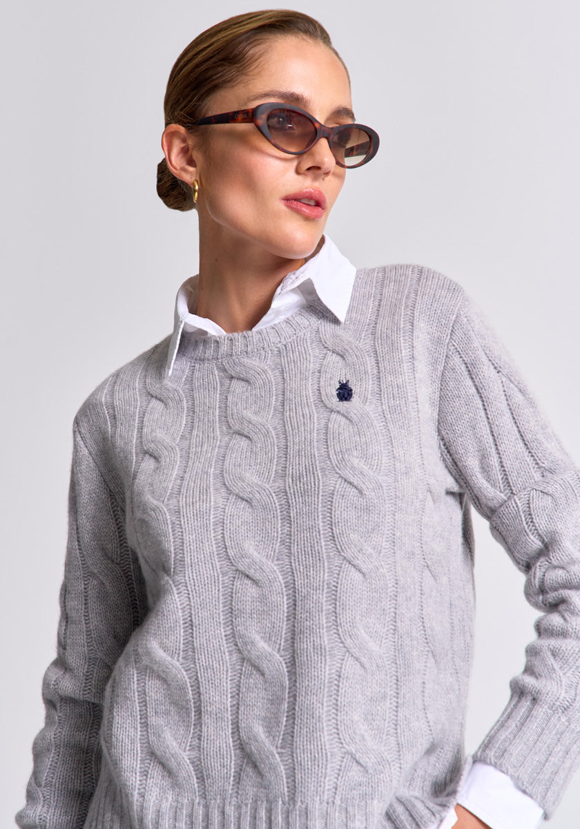 A woman wears brown sunglasses, a white collared shirt, and a light gray Princess Goes Hollywood wool-cashmere Zopfstrick pullover with a small embroidered frog emblem. Her hair is slicked back, she wears gold hoop earrings, and stands confidently against a plain background.
