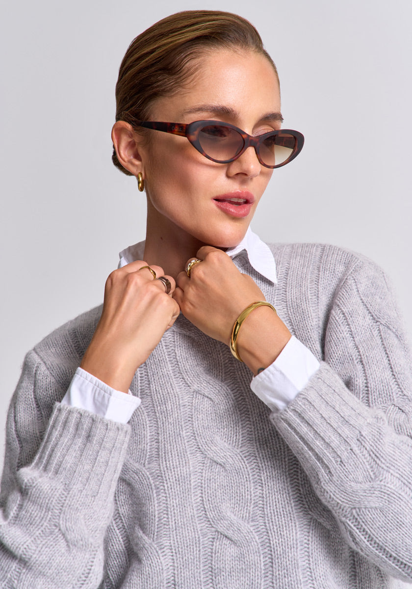 A woman in cat-eye sunglasses, gold hoop earrings, and a Princess Goes Hollywood Wolle-Kaschmir-Mix Pullover mit Zopfstrick poses confidently. Her sleek hair and chic gray sweater over a white shirt create a stylish look against a plain background.