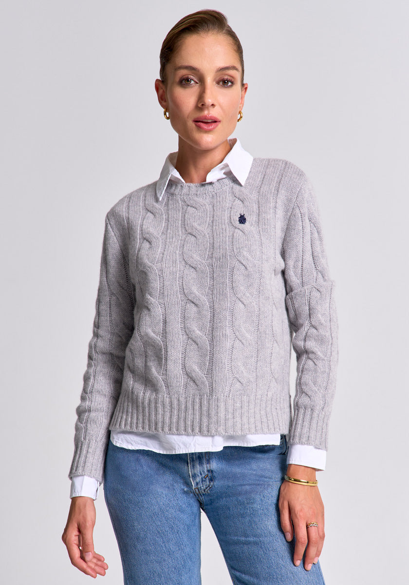 A woman with slicked-back hair wears a light gray Princess Goes Hollywood wool-cashmere blend pullover over a white collared shirt and blue jeans, accessorized with gold hoop earrings and a bracelet, standing before a plain light gray background.