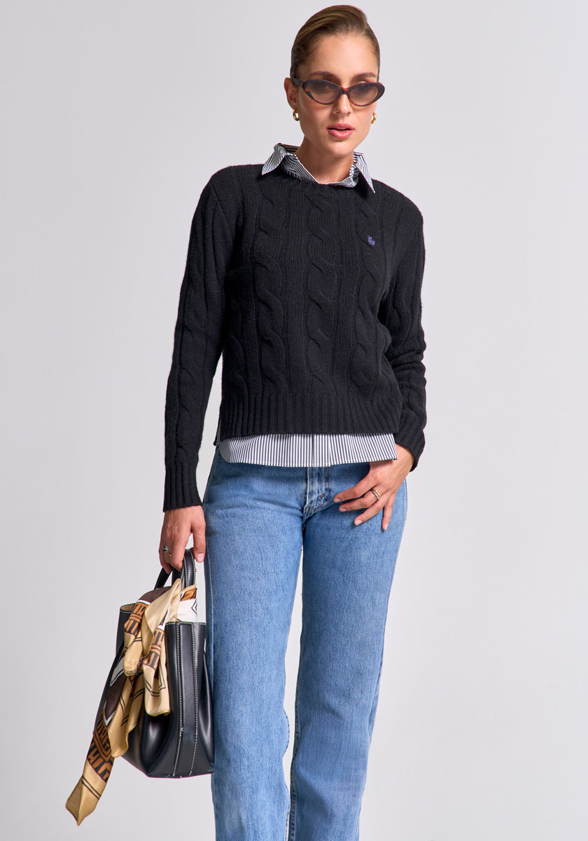 A woman wears brown sunglasses, a black Princess Goes Hollywood Zopfstrick Wolle-Kaschmir-Mix Pullover over a striped collared shirt, and blue jeans. She stands confidently with a black handbag and patterned scarf against a gray background.
