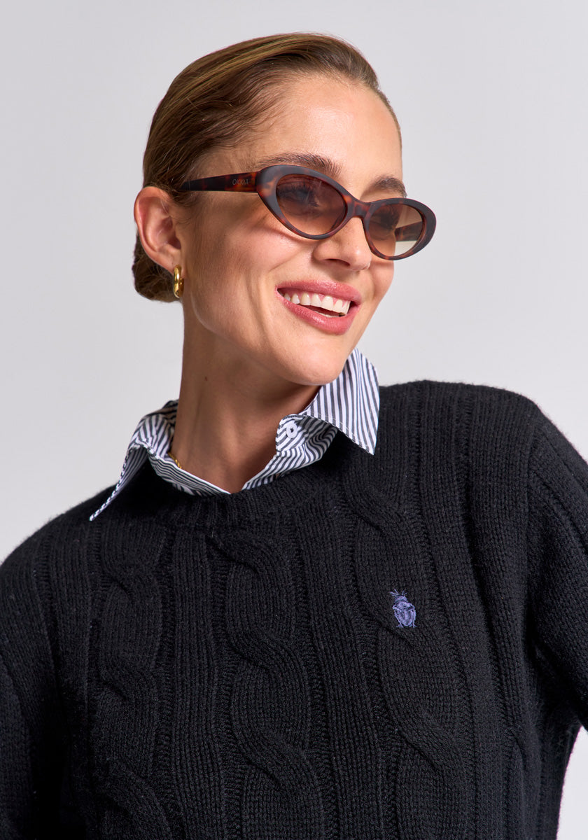 A smiling woman wears brown oval sunglasses, gold hoop earrings, and a Princess Goes Hollywood Regular Fit Zopfstrick Wolle-Kaschmir-Mix Pullover with crest logo over a blue-striped white collared shirt. Her slicked-back brown hair contrasts a light gray background.