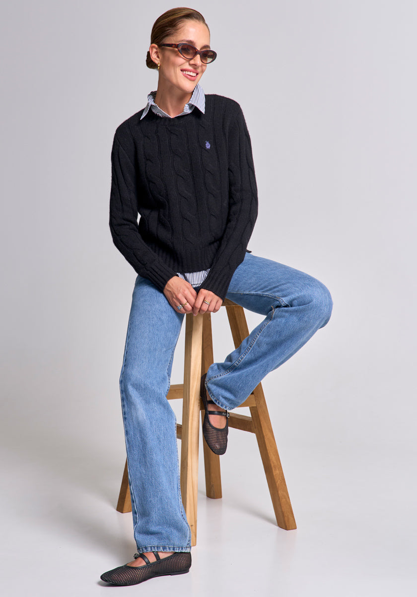 A woman with slicked-back hair, sunglasses, and small earrings sits on a wooden stool. She wears a Princess Goes Hollywood schwarzer Wolle-Kaschmir-Mix Pullover im Zopfstrick über einem Hemd, blue jeans, and dark flats.