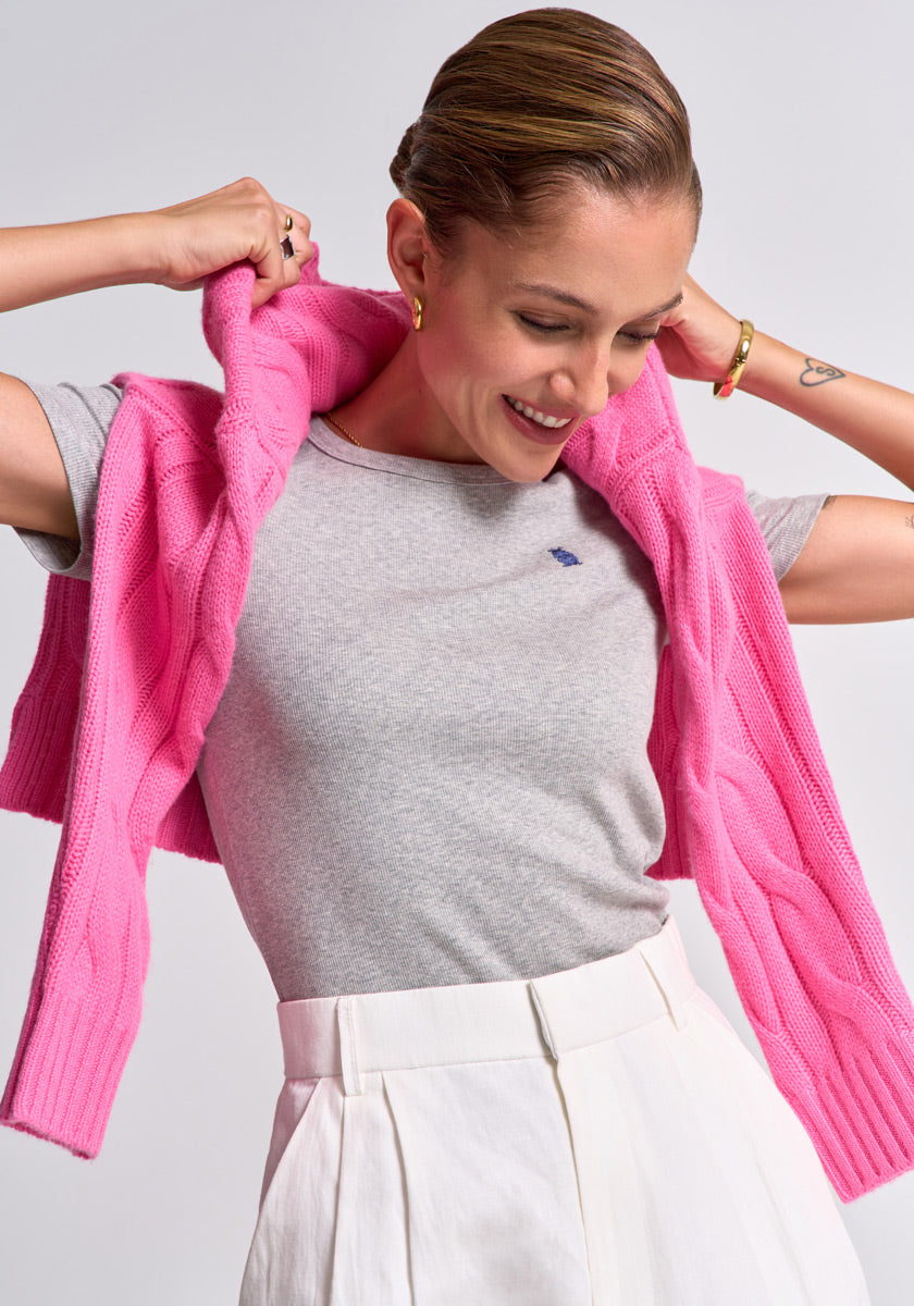 A smiling woman with slicked-back hair wears a grey V-Ausschnitt t-shirt tucked into high-waisted white pants. She drapes a bright pink Princess Goes Hollywood Zopfstrick Pullover Wolle-Kaschmir over her shoulders, accessorizing with gold jewelry.