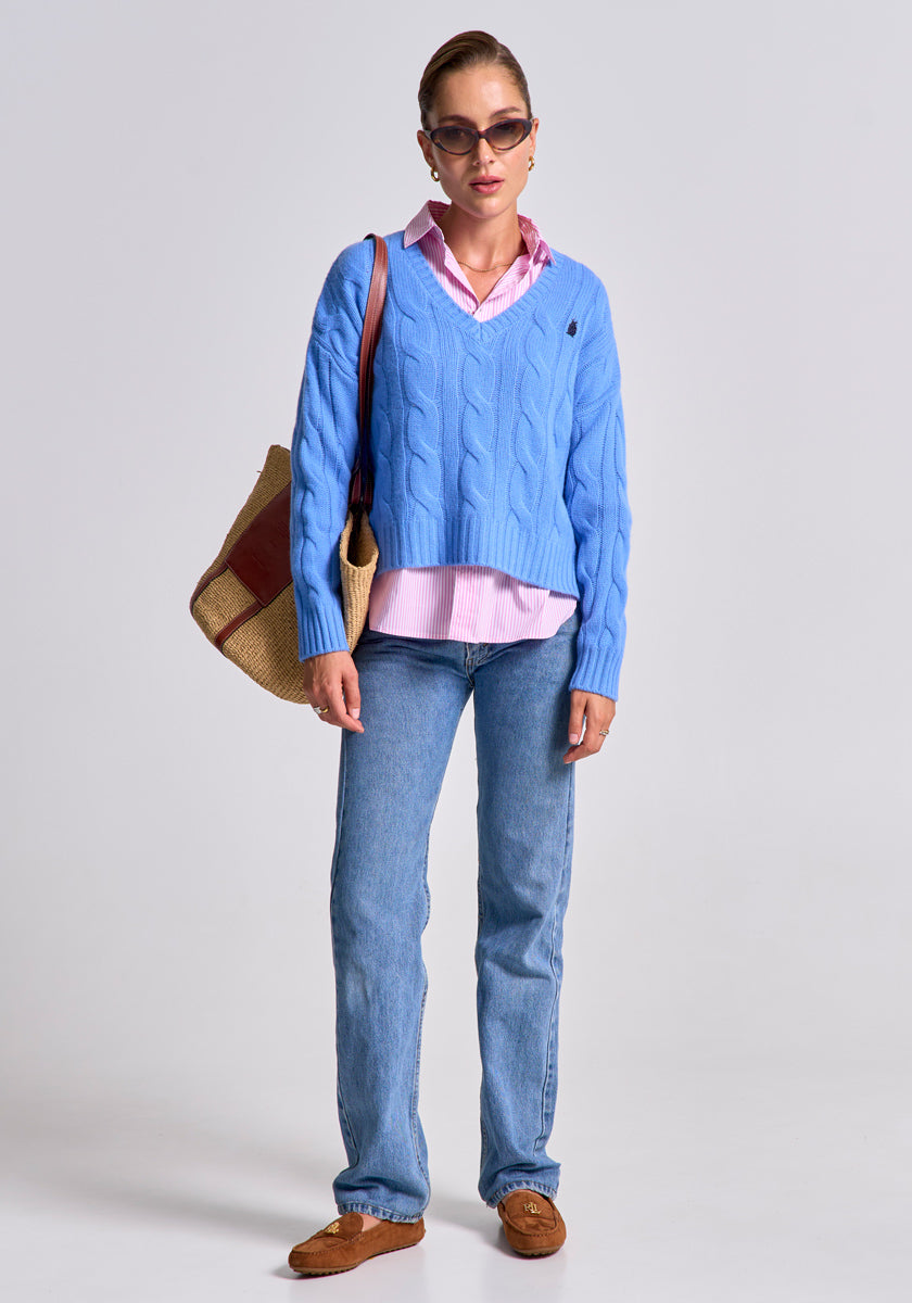 A woman stands against a plain background wearing a light blue Princess Goes Hollywood Zopfstrick Pullover aus Wolle-Kaschmir-Mix over a pink collared shirt, with blue jeans, brown loafers, sunglasses, and a red-accented woven tote bag.